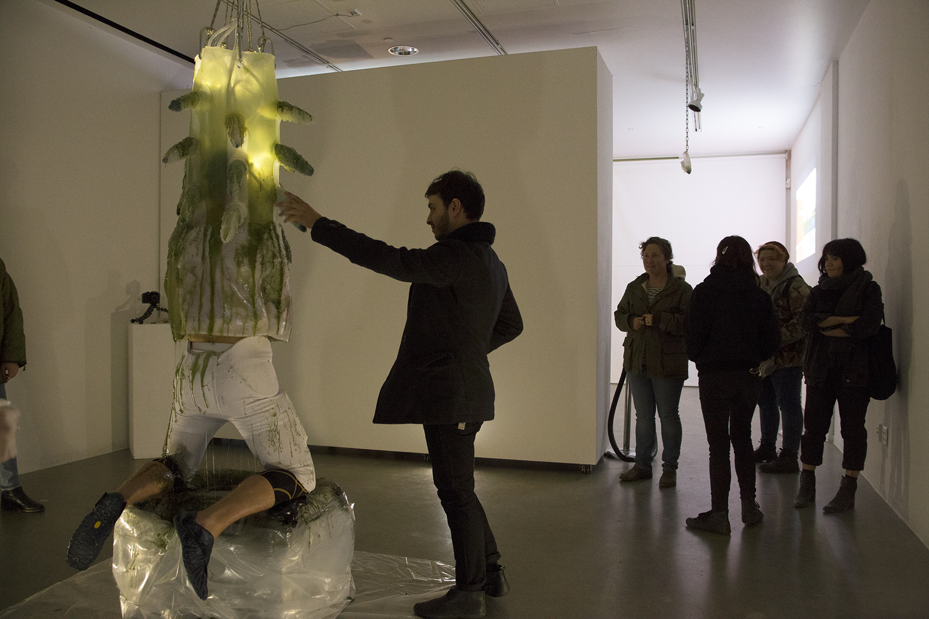 A man engulfed in a gelatenous form covered in tentacles knealing over a pool of green slime while a crowd of people watch